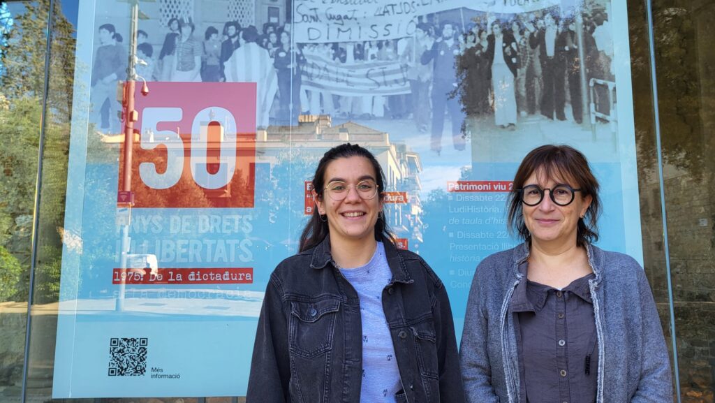 Esther Madrona i Alba Rodríguez davant del cartell promocional del programa al Museu del Monestir